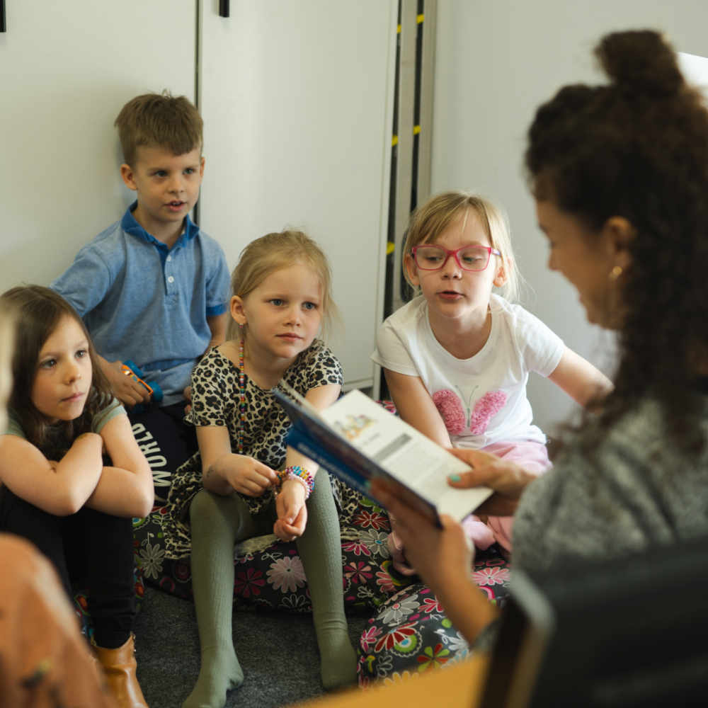 A woman reads a booklet to children sitting against the wall