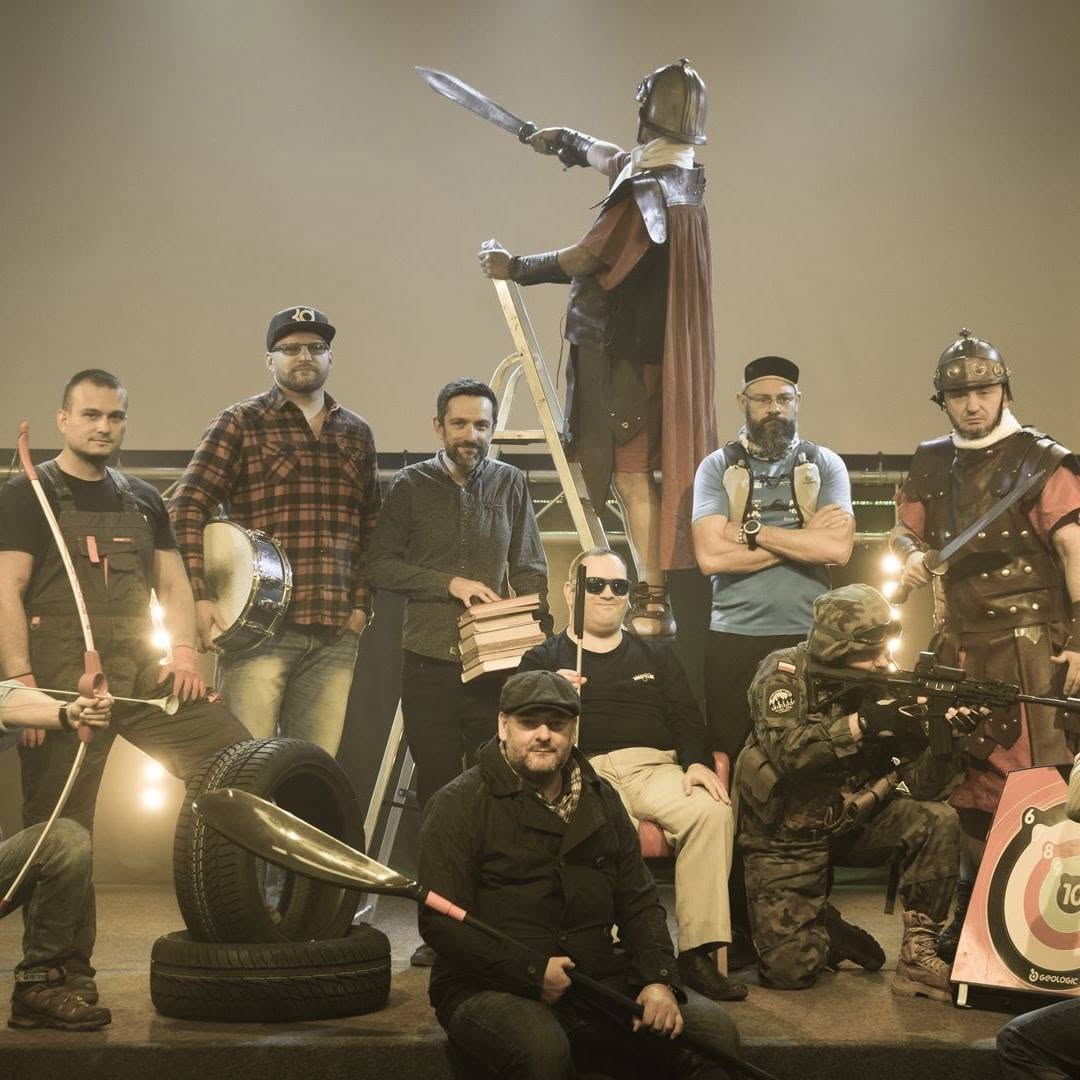 Men dressed as warriors, holding swords, a bow and a shield, pose among tyres and an archery target