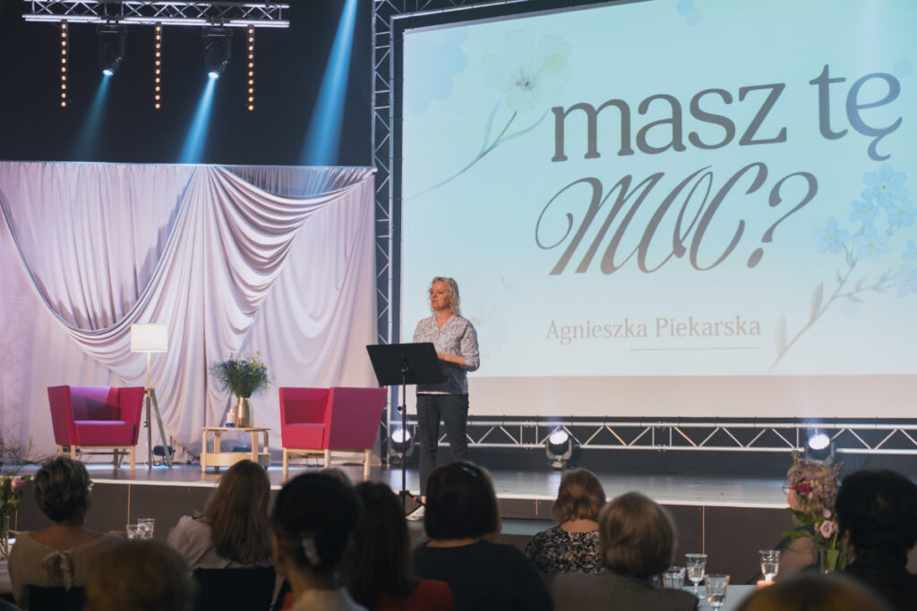 Kobieta przemawia na scenie podczas spotkania kobiet, za nią ekran z hasłem „Masz tę moc?”