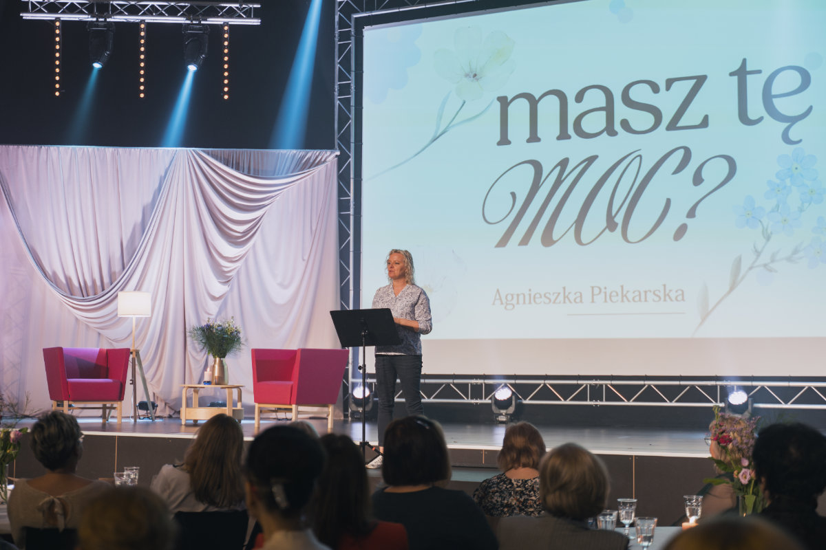 A woman speaks on stage at a women’s gathering, with the title “Masz tę moc?” on the screen behind her